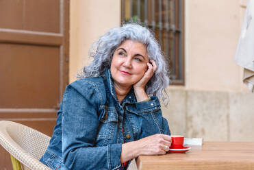 Cheerful elderly female in denim jacket with curly gray hair sitting at table with cup of hot aromatic coffee and looking away while resting with her chin resting upon her hand in outdoor cafe - ADSF45148