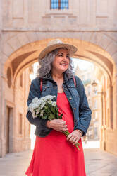 Cheerful mature female in denim jacket and red dress with bouquet of flowers and straw hat smiling and looking away while walking on city street - ADSF45151
