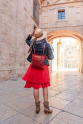 Back view full body of anonymous female tourist with backpack wearing red dress and hat standing on paved walkway near historic buildings during sightseeing trip - ADSF45152