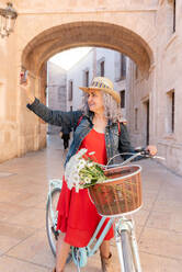 Side view of full body of cheerful senior female taking photo on smartphone while standing near vintage bicycle with basket of flowers on paved street - ADSF45153