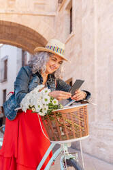 Happy mature female in elegant outfit and straw hat smiling and messaging on mobile phone while leaning on bicycle with basket of white flowers on street - ADSF45154