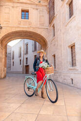 Full body of cheerful senior female in stylish outfit and straw hat smiling and looking away near bicycle with basket of flowers on paved street - ADSF45155