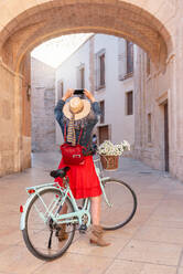 Back view of full body of cheerful senior female taking photo on smartphone while standing near vintage bicycle with basket of flowers on paved street - ADSF45157