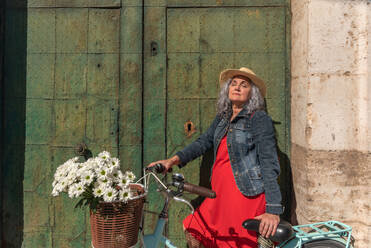 Dreamy senior female in casual outfit and straw hat standing with closed eyes near vintage bicycle with bouquet of flowers in wicker basket on sunny street - ADSF45160