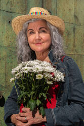 Cheerful mature female in denim jacket and red dress with bouquet of flowers and straw hat smiling and looking at camera while standing on city street - ADSF45161