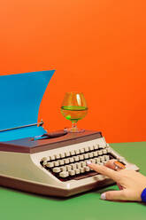 Crop anonymous person at table with glass of wine typing on old typewriter with blue carton paper over green two colored background - ADSF45192