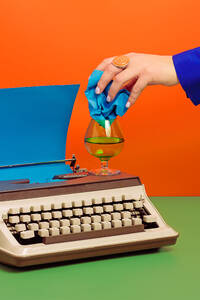 Crop woman with typewriter and blue paper putting crumpled paper into glass cup with drink all on green surface against orange background - ADSF45193