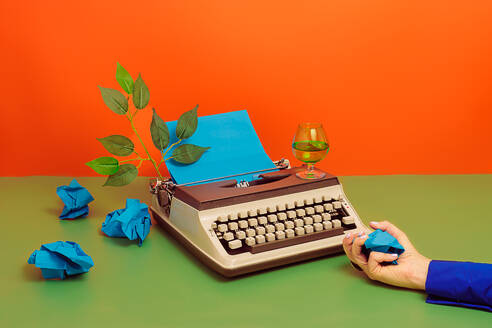 Crop hand of person in blue shirt holding crumpled paper at old typewriter with keyboard and cup of wine with green branch over multicolored surface in studio - ADSF45194