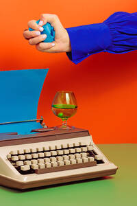 Crop woman with typewriter and blue paper putting crumpled paper on glass cup with drink all on green surface against orange background - ADSF45195