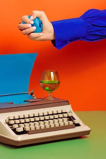 Crop woman with typewriter and blue paper putting crumpled paper on glass cup with drink all on green surface against orange background - ADSF45195