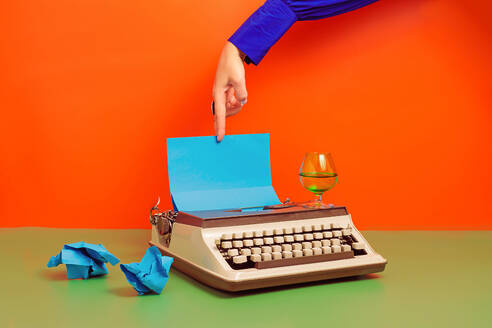 Crop anonymous person with hand over typewriter with blue paper and cup of drink and squeezed paper over smooth green surface against orange background - ADSF45200