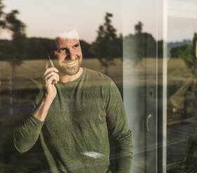 Lächelnder Mann, der durch ein Glas hindurch mit seinem Handy telefoniert - UUF29403