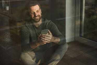 Glücklicher Mann mit Mobiltelefon durch Glas gesehen - UUF29407