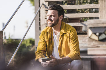 Glücklicher Mann sitzt mit Smartphone an einem sonnigen Tag - UUF29424