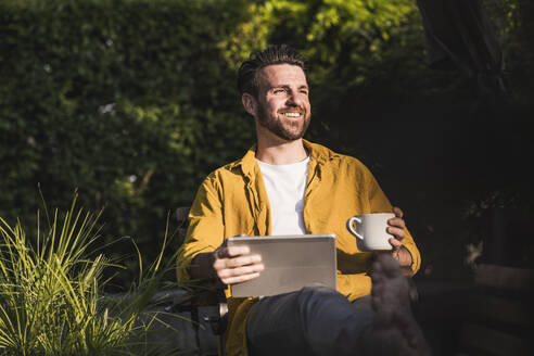 Glücklicher Mann sitzt mit Tablet-PC und Kaffeetasse - UUF29430