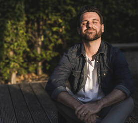 Smiling man with hands clasped and eyes closed sitting on porch - UUF29448