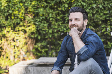 Smiling man with hand on chin sitting in front of plants - UUF29450