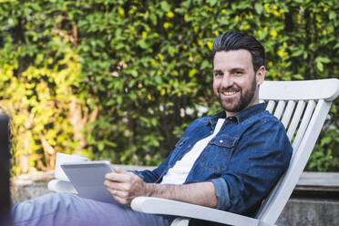 Lächelnder Mann sitzt mit Tablet-PC und Kaffeetasse auf einem Stuhl - UUF29454