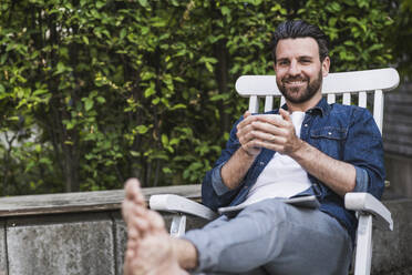 Smiling man sitting with coffee cup on chair - UUF29455