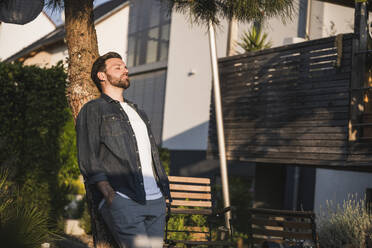 Man with eyes closed leaning on tree trunk in front of house - UUF29466