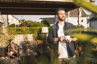 Glücklicher Mann mit Kaffeetasse auf der Terrasse stehend - UUF29469