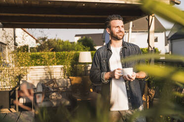 Happy man standing with coffee cup on terrace - UUF29469