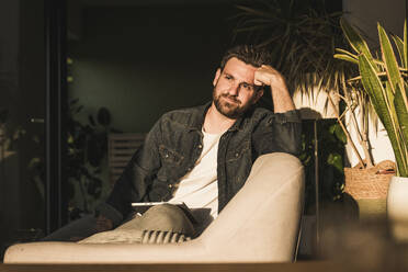 Thoughtful man sitting on sofa with tablet PC at home - UUF29475