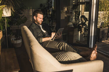 Smiling man using tablet PC on sofa in living room at home - UUF29481