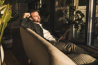 Thoughtful man sitting with tablet PC on sofa in living room at home - UUF29482