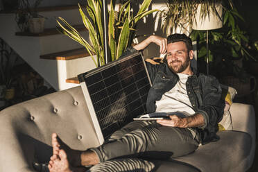 Glücklicher Freiberufler sitzt mit Tablet-PC und Solarpanel auf dem Sofa - UUF29487