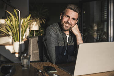 Happy thoughtful freelancer sitting with laptop at desk - UUF29494