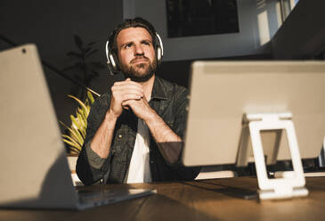 Thoughtful freelancer wearing headset sitting at desk - UUF29500
