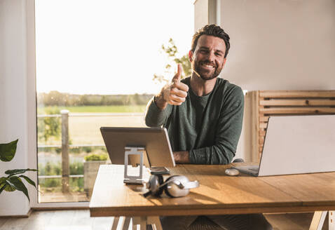 Happy freelancer showing thumbs up gesture at desk - UUF29503
