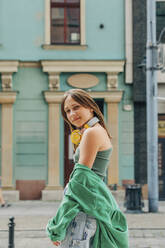 Smiling girl with wireless headphones standing in front of building - VSNF01202