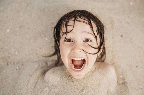 Fröhlicher Junge mit Sand bedeckt, der am Strand Spaß hat - ANAF01750
