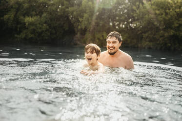 Glücklicher Vater und Sohn beim Schwimmen im Fluss an einem sonnigen Tag - ANAF01758