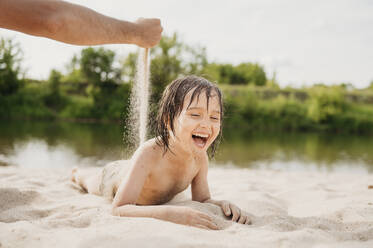 Fröhlicher Junge mit Vater, der Sand fallen lässt und sich am Strand vergnügt - ANAF01759