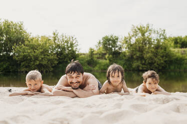 Father lying on sand with children on riverbank - ANAF01760