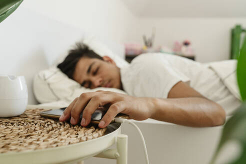 Müder Mann hält Smartphone auf Beistelltisch in der Nähe des Bettes - OSF01866