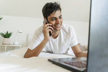 Lächelnder Mann liegt auf dem Bett und spricht mit seinem Smartphone zu Hause - OSF01871
