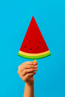 Crop unrecognizable person demonstrating bright paper watermelon popsicle against blue background in studio - ADSF45201