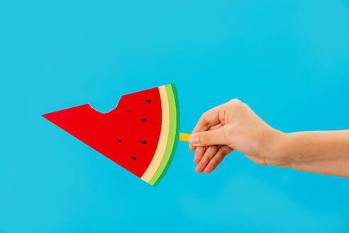 Crop unrecognizable person demonstrating bright paper watermelon with a bite out popsicle against blue background in studio - ADSF45203