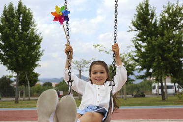 Girl with pinwheel toy swinging on swing - SYEF00487