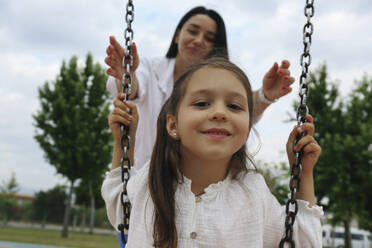 Smiling mother with daughter sitting on swing in park - SYEF00490