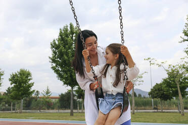 Happy mother spending time with daughter sitting on swing in park - SYEF00493