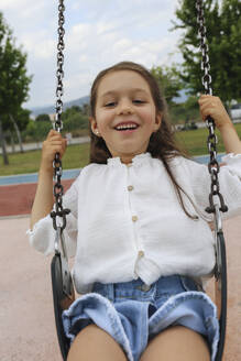 Glückliches Mädchen sitzt auf einer Schaukel auf dem Spielplatz - SYEF00494