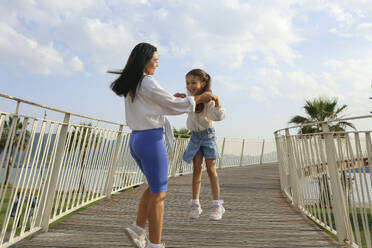 Happy mother holding hands with daughter jumping on bridge - SYEF00500
