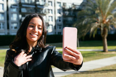 Eine fröhliche hispanische Frau in einer Lederjacke macht ein Selfie mit ihrem Handy, während sie bei Tageslicht auf einer Straße in der Stadt steht - ADSF45217