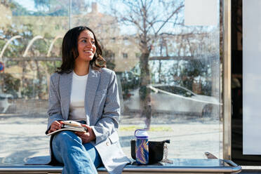 Positive young Hispanic female student sitting cross legged on bench with books water bottle at city bus stop with glass walls and smiling while looking away in daylight - ADSF45220