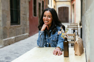 Positive young Hispanic female with hand at chin leaning on table with handbag and smiling while looking at camera in street cafe - ADSF45222
